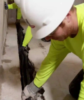 a man wearing a hard hat is kneeling down and working with a HydraDry basement waterproofing system