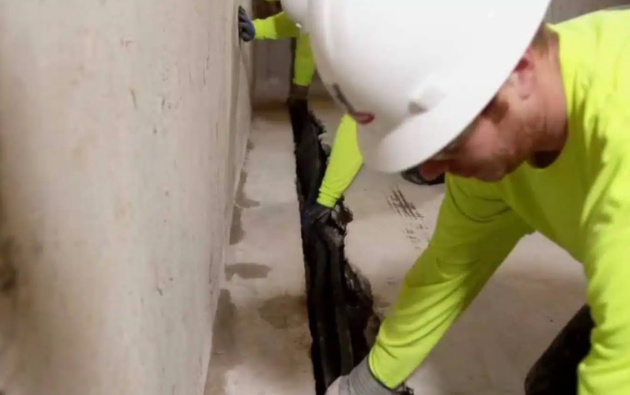 a man wearing a hard hat is kneeling down and working with a HydraDry basement waterproofing system